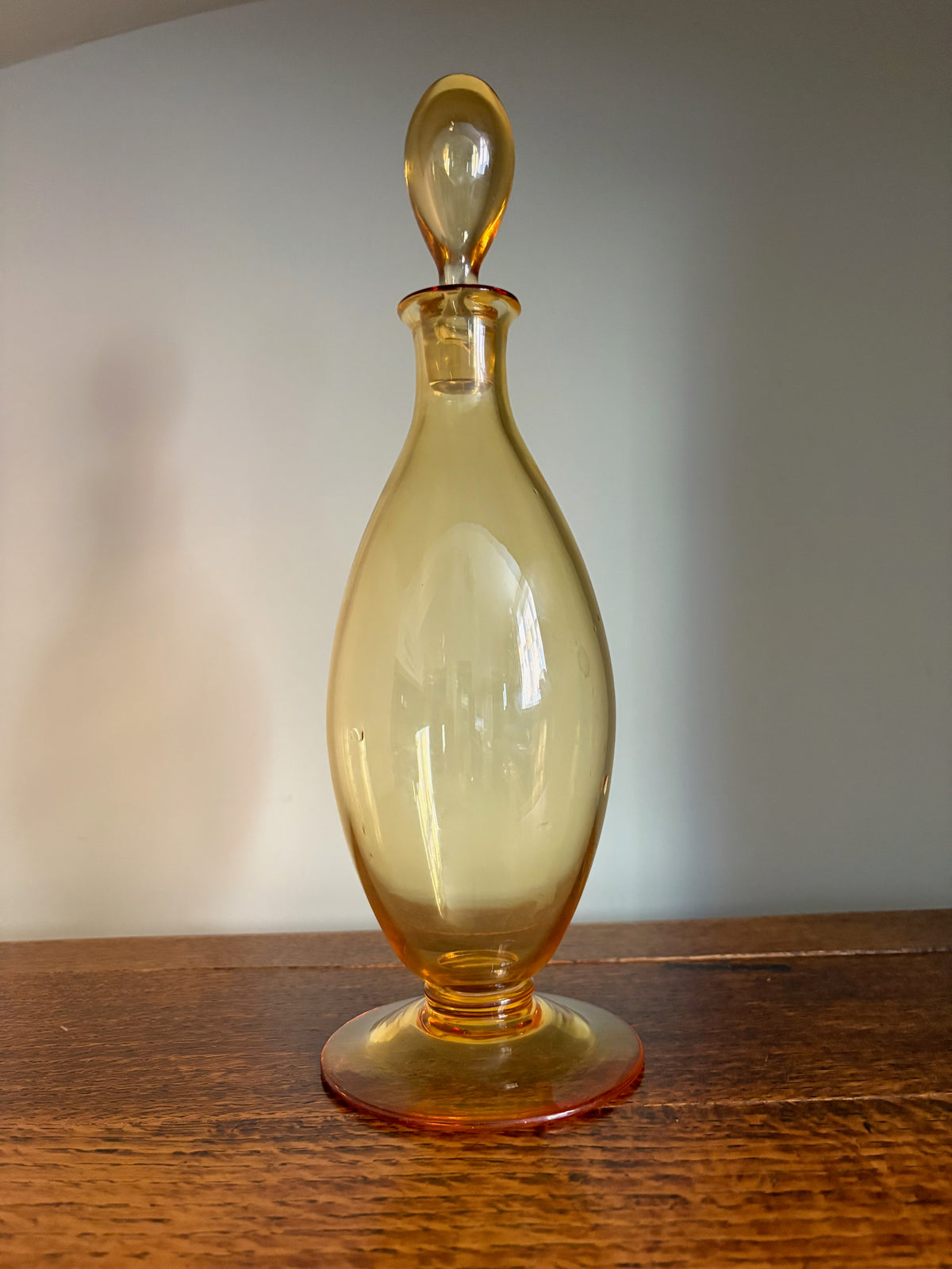 A vintage Whitefriars amber glass decanter with a matching stopper, resting on a oak wood tabletop against a plain cream wall.