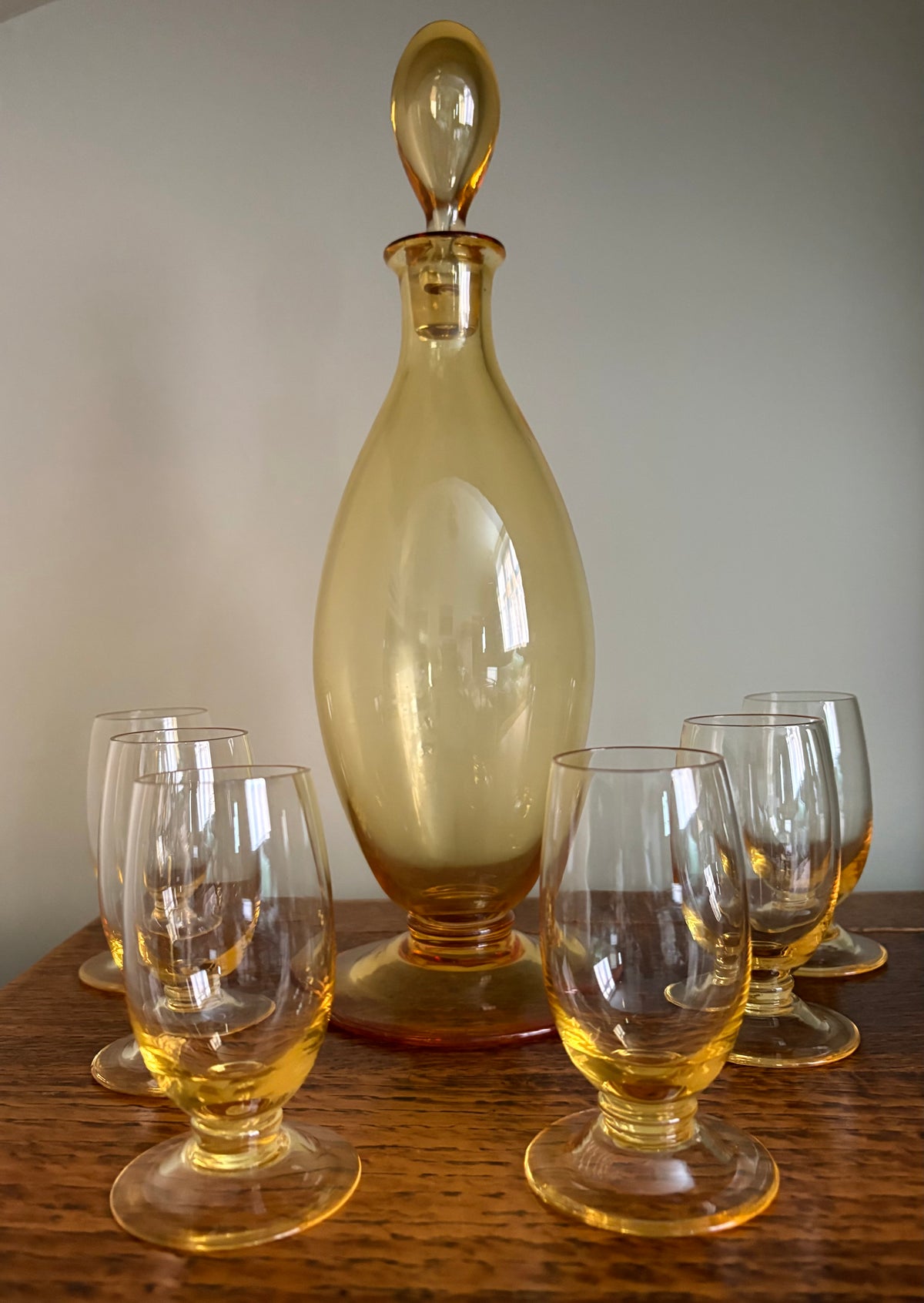 A vintage Whitefriars amber glass decanter with a stopper, displayed on a wooden table withsixvmatching amber-tinted glasses against a neutral cream background.