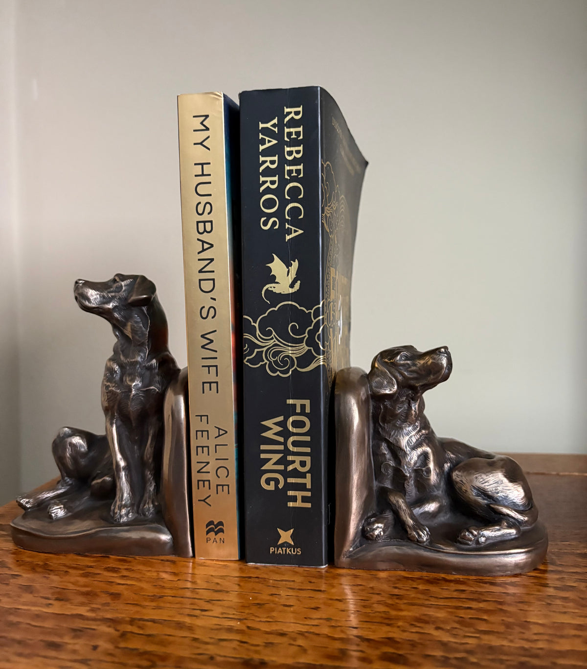 Pair of Genesis Heredities cast resin bronze effect bookends, modelled as Labrador dogs and shaped one seated and one lying down holding up two books on a wooden table against a plain wall.