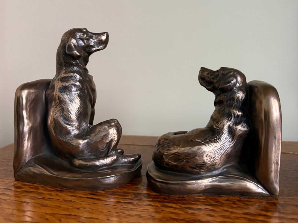 Pair of Genesis Heredities cast resin bronze effect bookends, modelled as Labrador dogs and shaped one seated and one lying down, on a wooden table against a plain light gray wall.