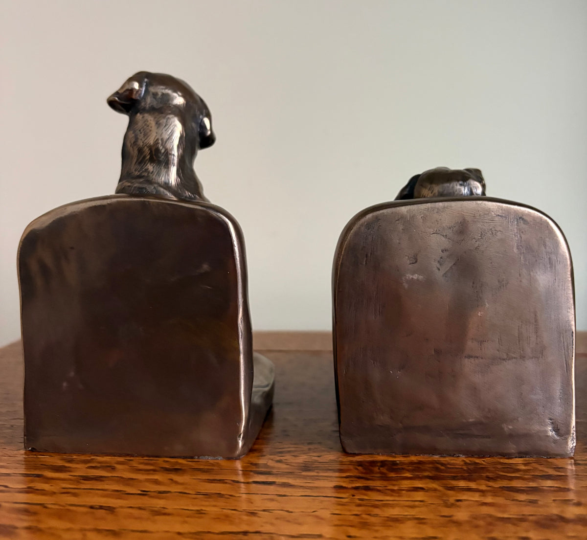 Pair of Genesis Heredities cast resin bronze effect bookends, modelled as Labrador dogs and shaped one seated and one lying down, shown from the back, resting on a wooden table against a plain light gray wall.