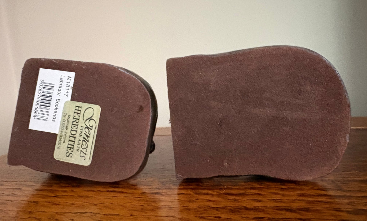 The underside of pair of Genesis Heredities cast resin bronze effect bookends, showing the brown material bases, and one showing a barcode sticker and the maker label, resting on a wooden table against a plain wall.