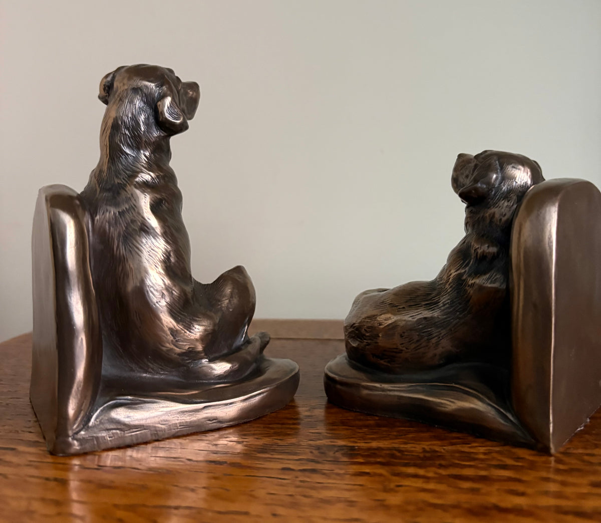 Back view of a pair of Genesis Heredities cast resin bronze effect bookends, modelled as Labrador dogs and shaped one seated and one lying down on a wooden table against a plain light wall.